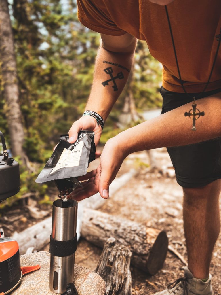 A man pours coffee beans into a grinder perched onto a wooden stump in the woods. 